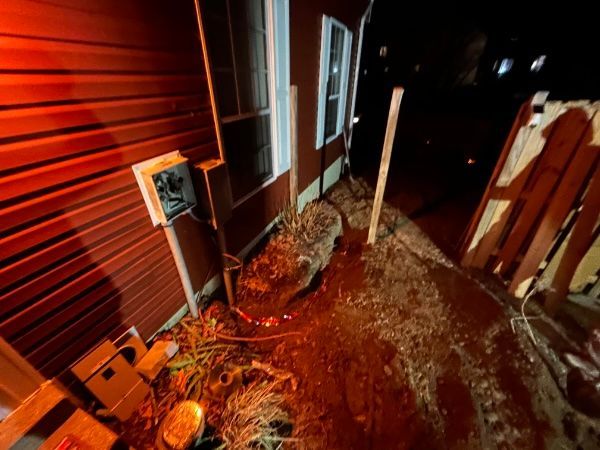 Red-sided building at night with a small electrical box, window, and debris-covered ground; wooden fence visible.
