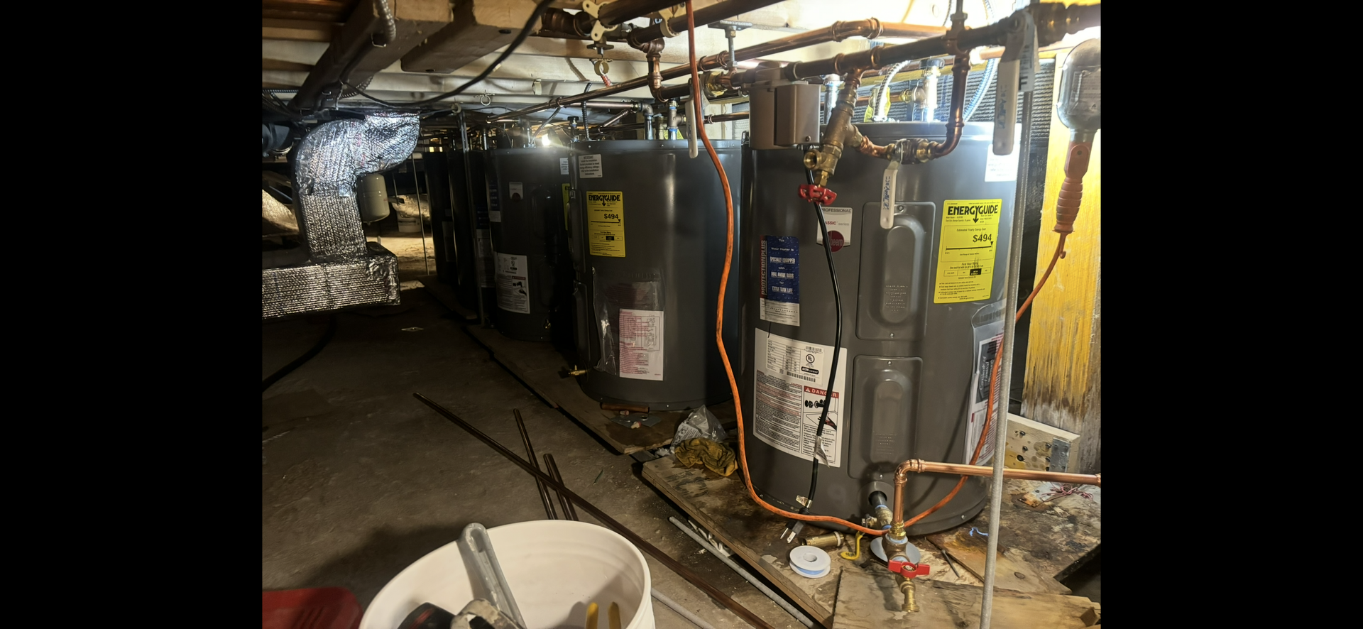 A row of gray water heaters in a dimly lit basement. Copper pipes and wiring are visible.