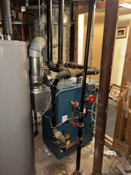 A boiler and water heater in a basement. Black pipes, silver chimney, blue boiler, light gray water heater.