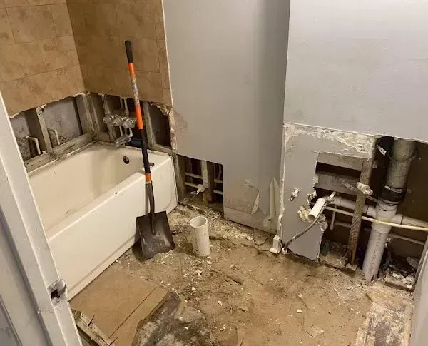 Bathroom renovation in progress, walls open to studs, bathtub, shovel, and debris on the floor.