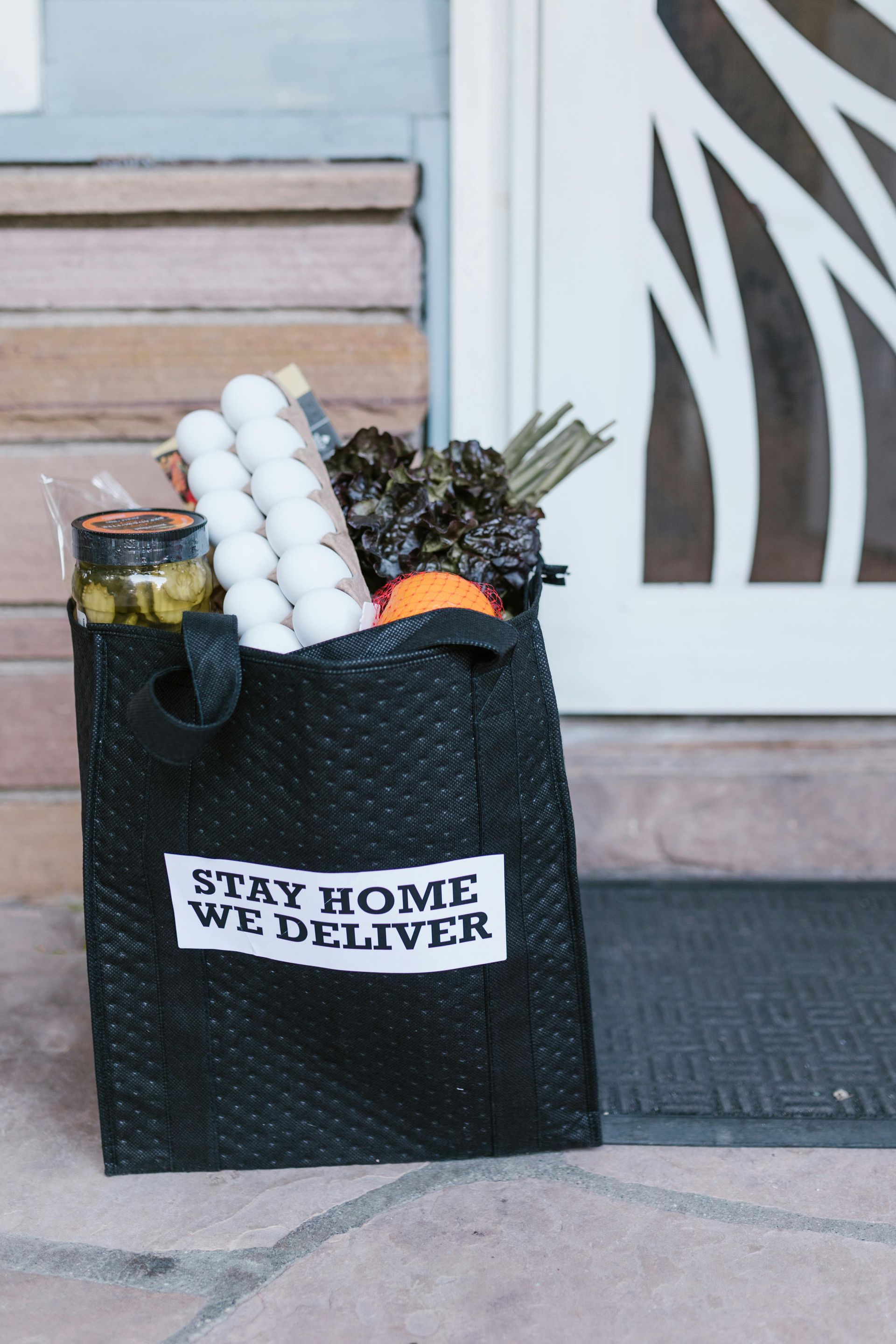Gig delivery bag with groceries on doorstep displaying 'Stay Home We Deliver' label