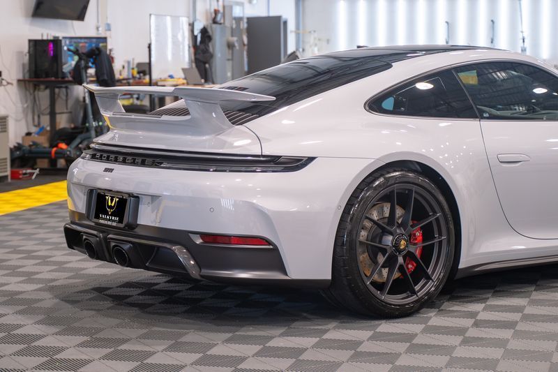 White Porsche sports car rear view with spoiler, parked in a garage with black tire rims.