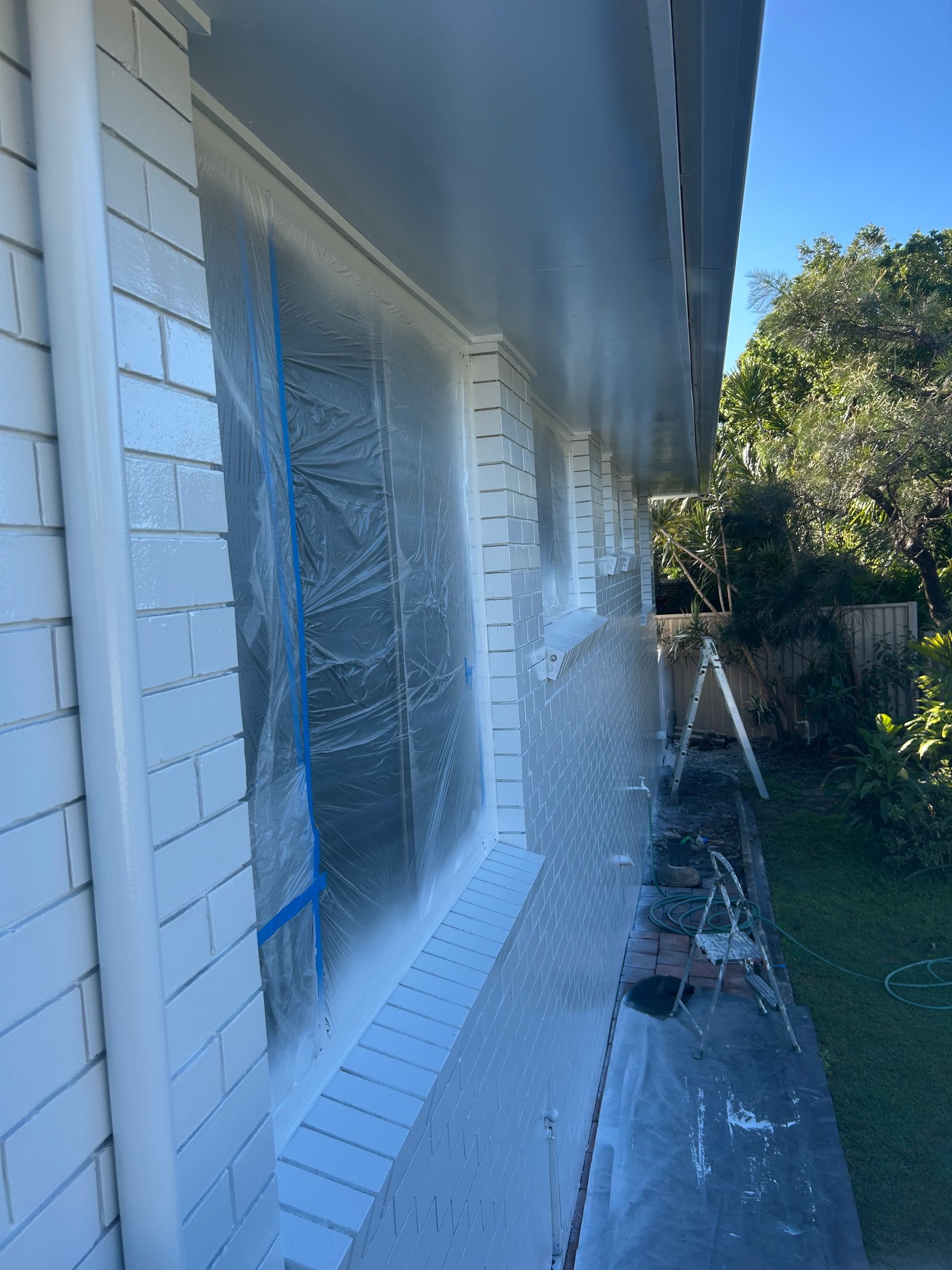 Exterior house painting in progress; white paint spraying on window trim and siding.
