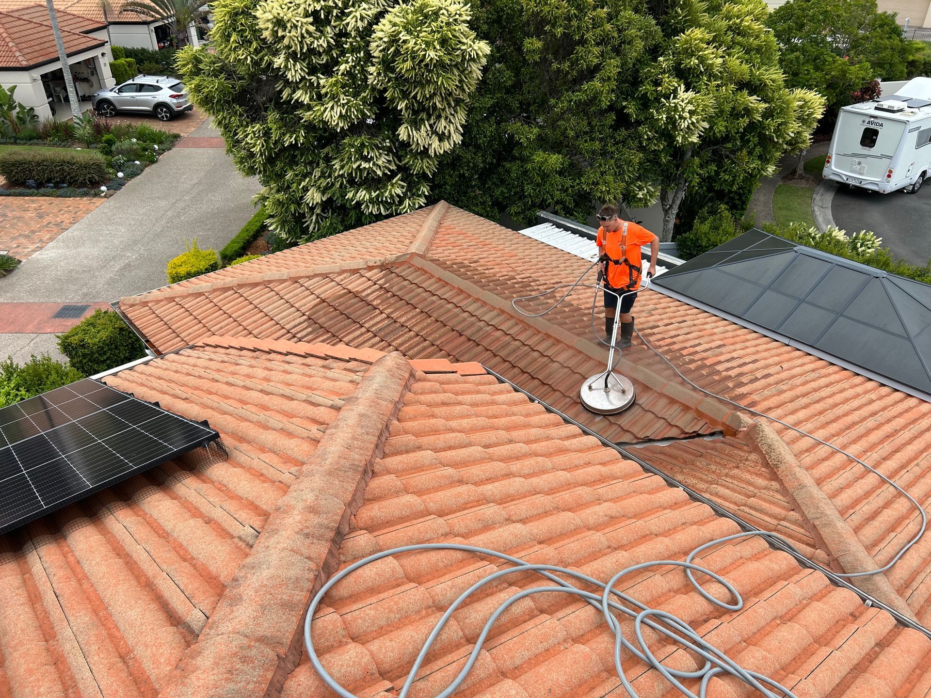 Person in orange shirt pressure washing a terracotta tiled roof. Rooftop setting, solar panels.