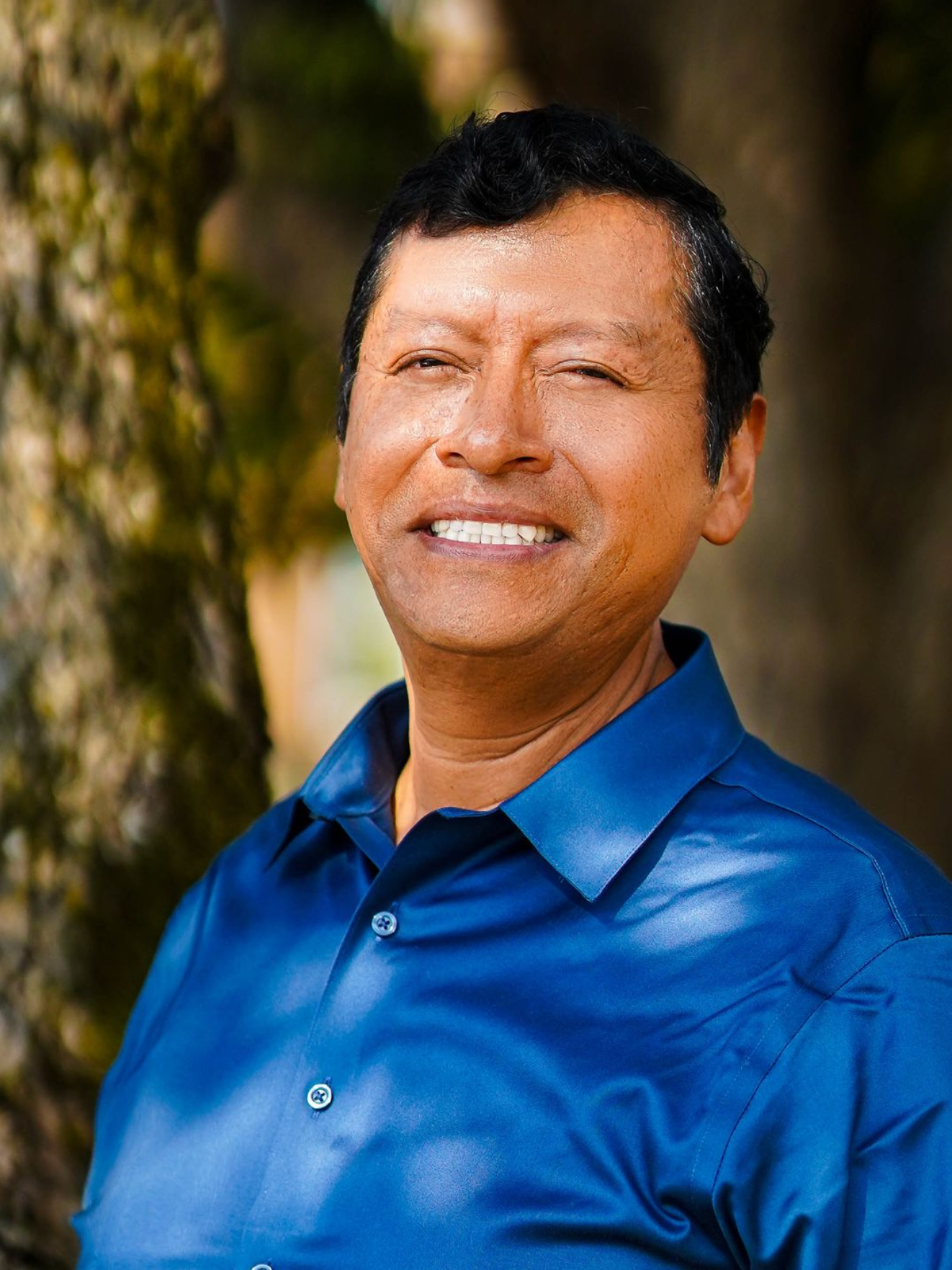 Man smiling, wearing a blue shirt, outdoors near a tree.