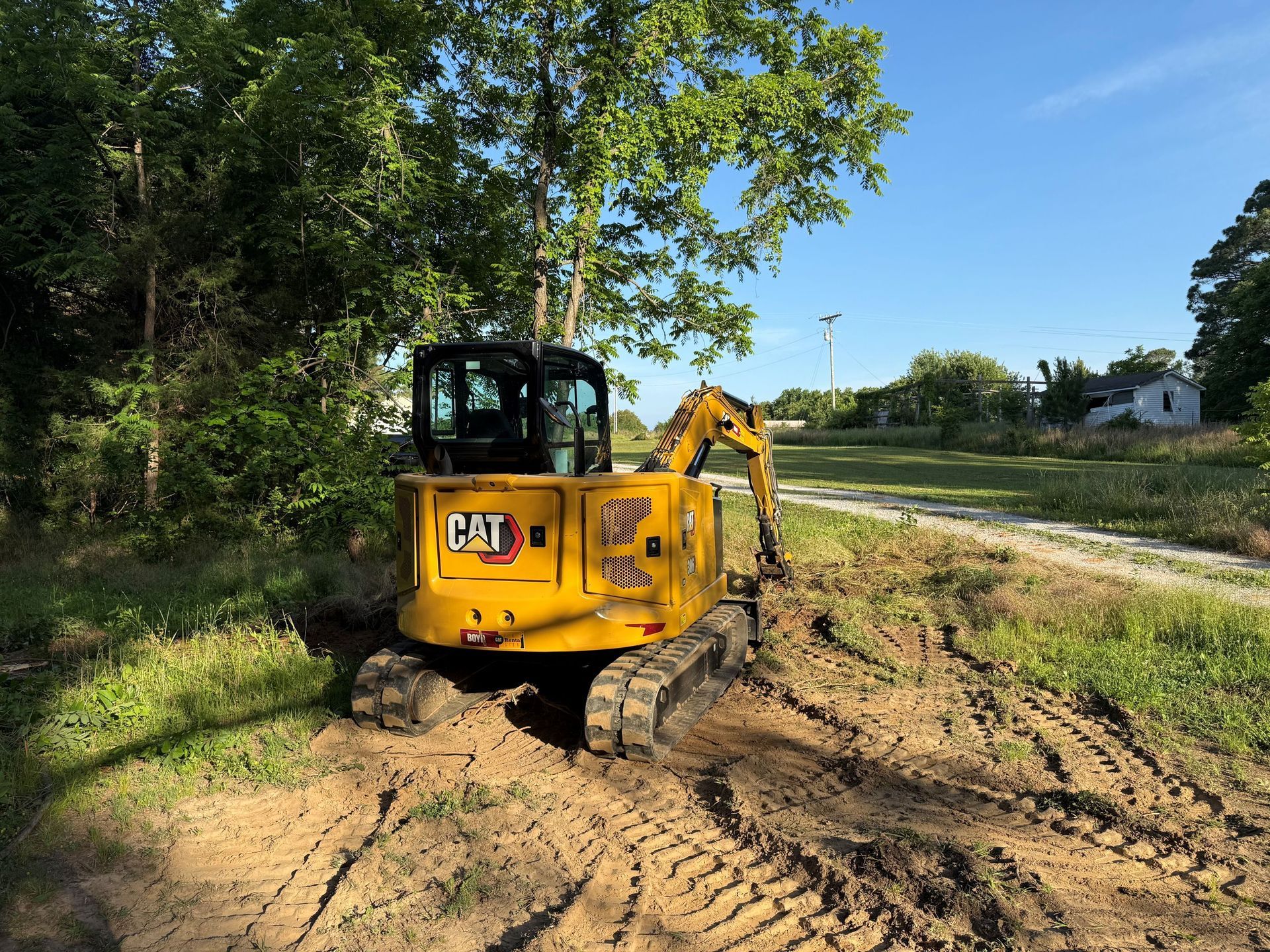 Yellow excavator clearing a dirt path beside trees and a road on a sunny day