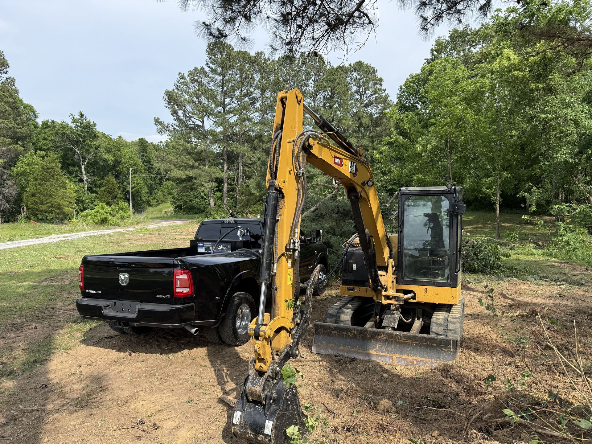 Yellow excavator beside a black pickup truck on a grassy wooded lot