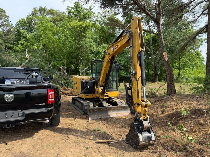 Black pickup and yellow excavator on a dirt path in a wooded area