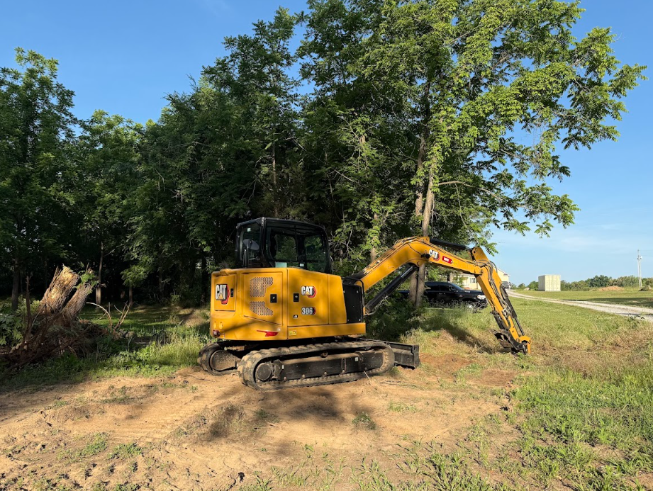 Yellow excavator parked on a dirt clearing beside trees and a grassy field