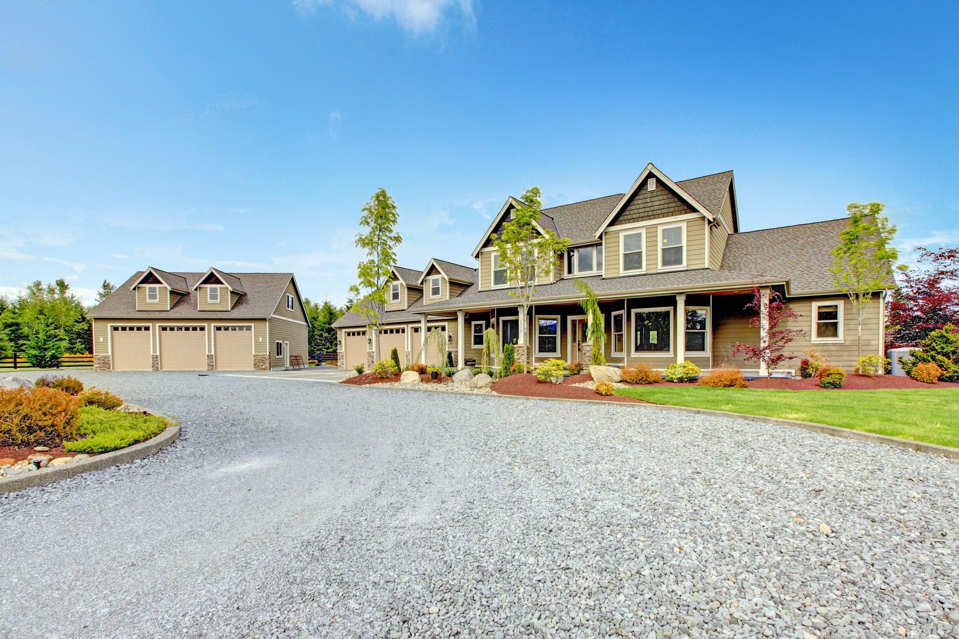 Large modern farmhouse with gravel driveway, manicured landscaping, and detached garage under a clear sky