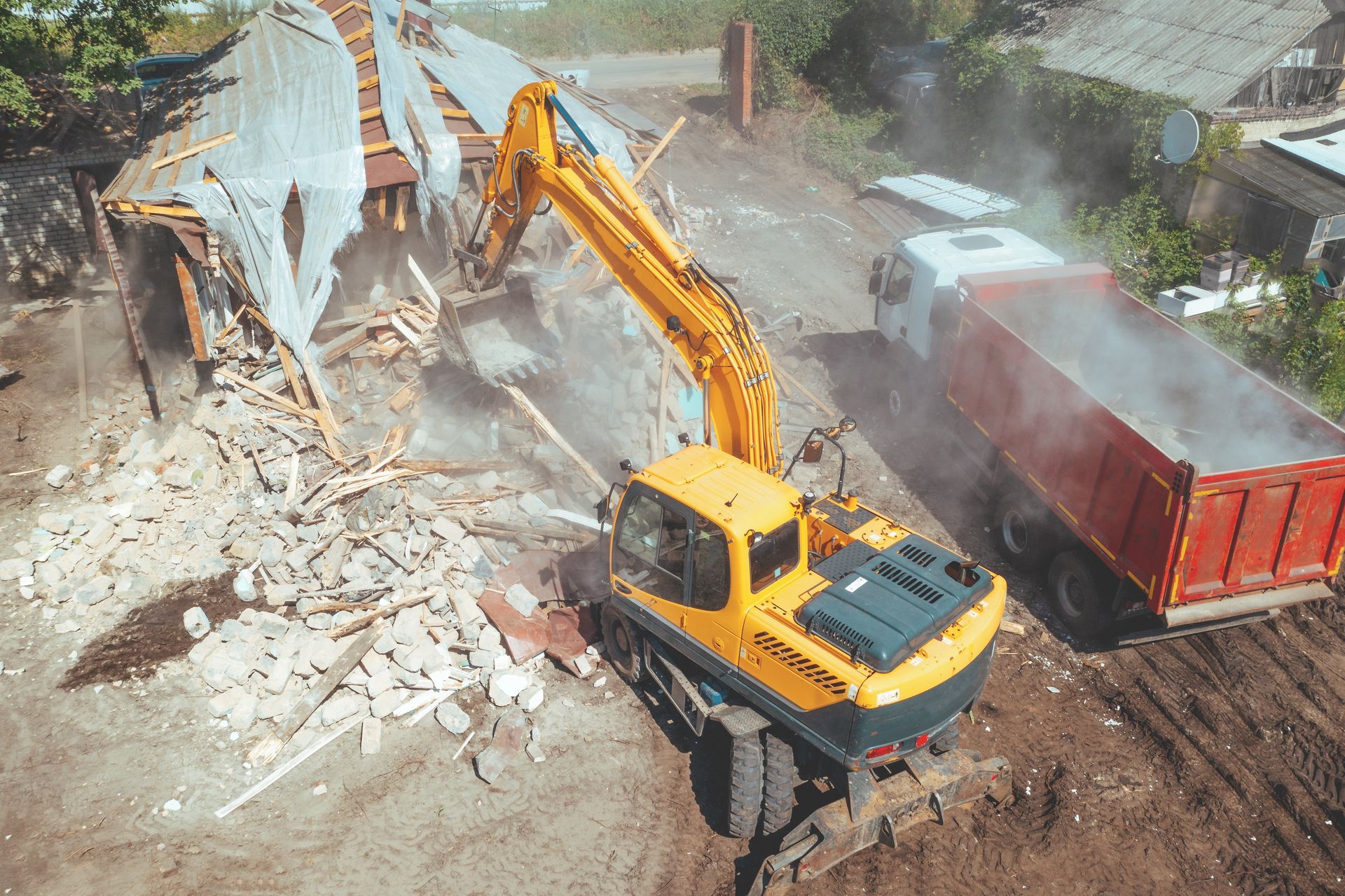 Excavator demolishing a building beside a red dump truck, with dust and debris on a construction site
