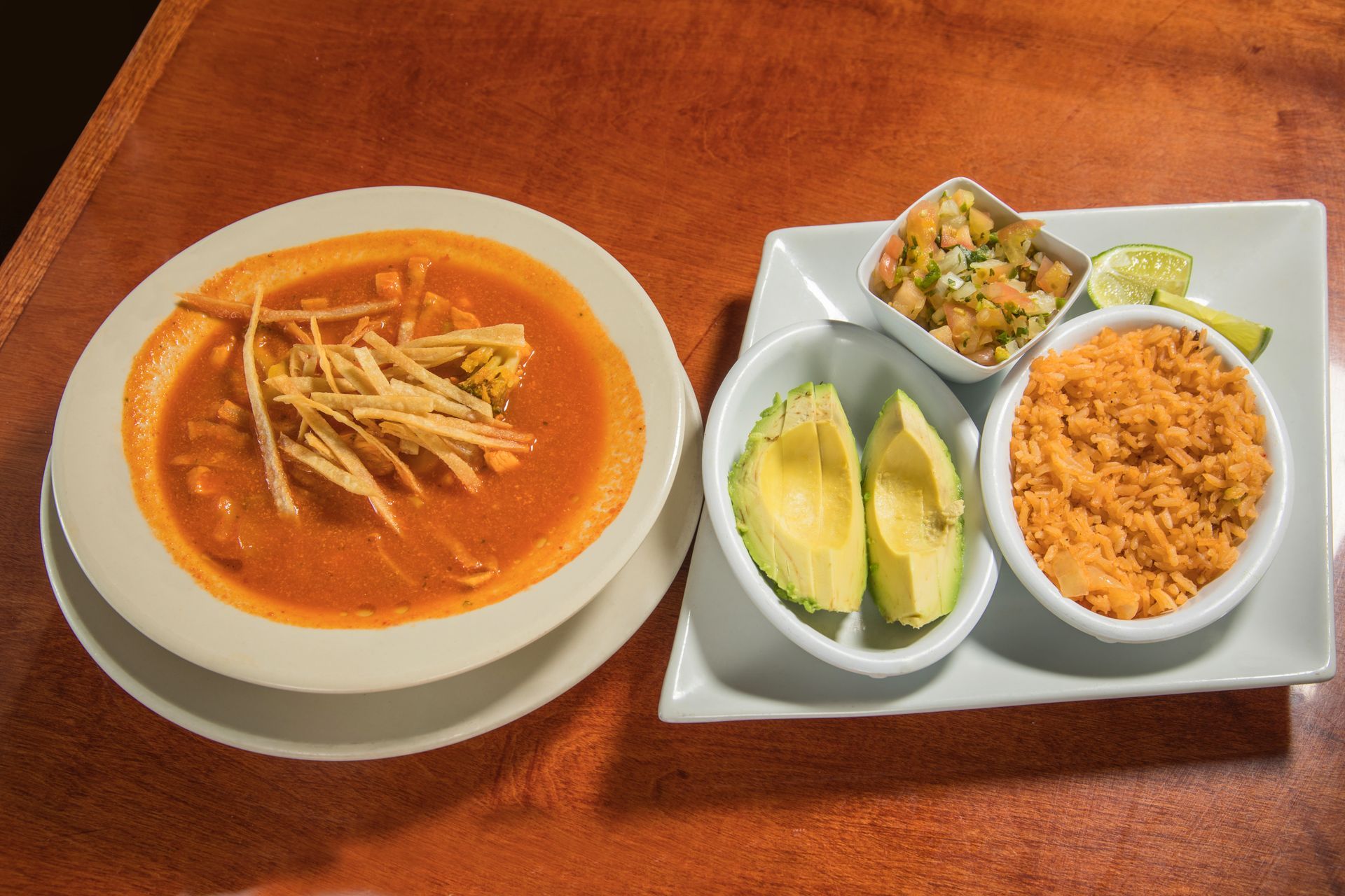 A bowl of soup next to a plate of rice and avocado.