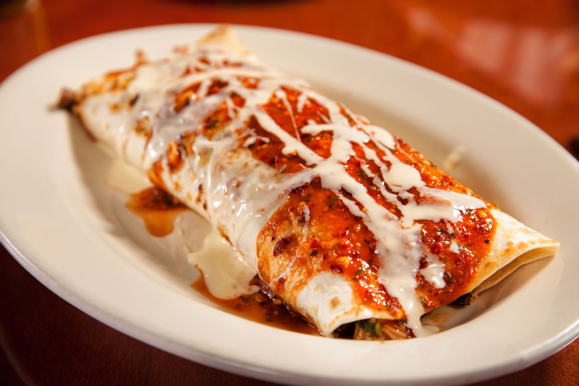 A burrito with sauce and cheese on a white plate on a table.