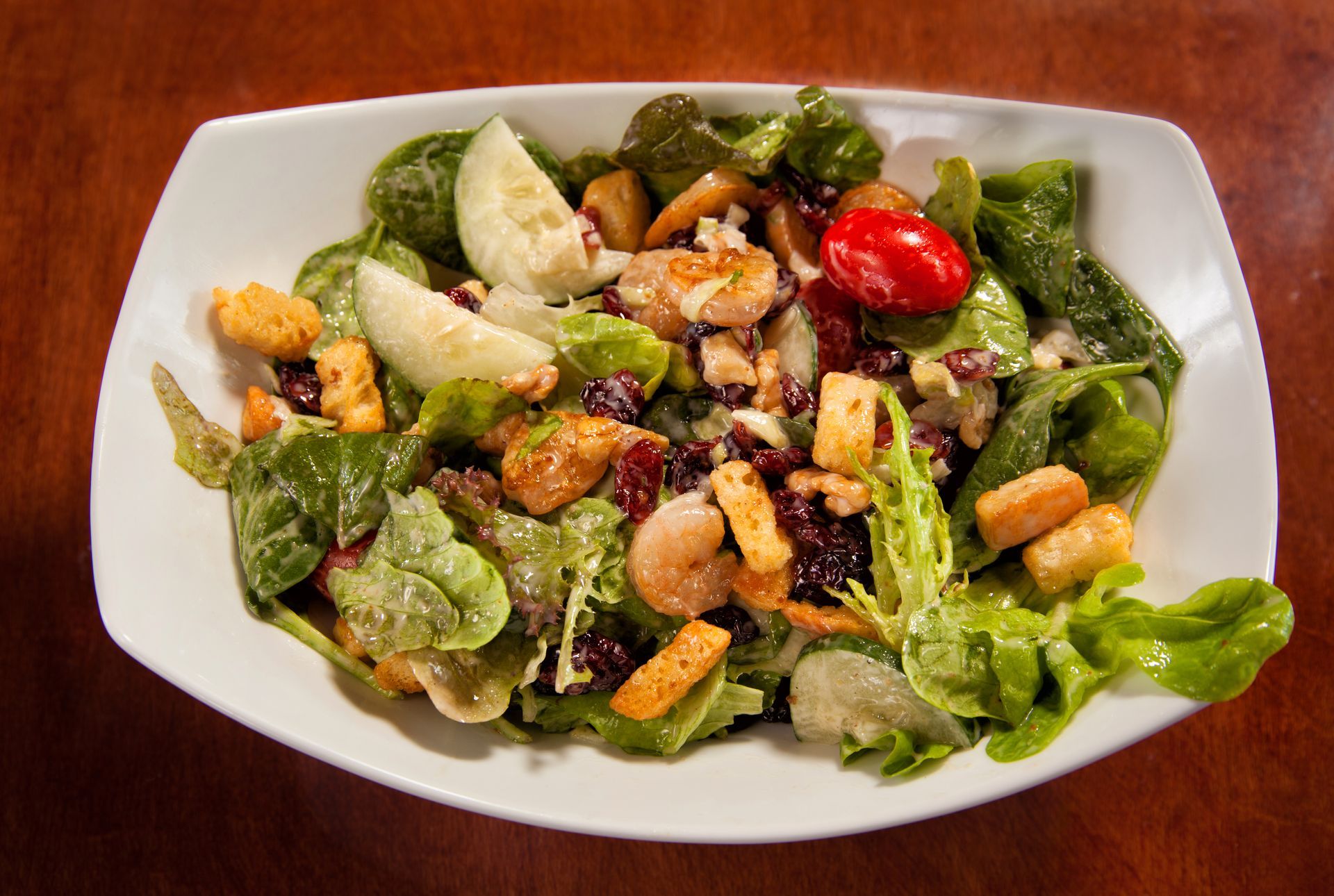 A salad with shrimp , lettuce , tomatoes and croutons on a white plate on a wooden table.