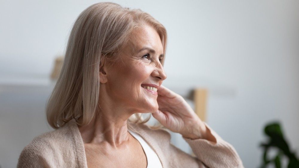 An Older Woman Is Smiling And Touching Her Face While Looking Out A Window — Mackay Skin Clinic In North Mackay, QLD