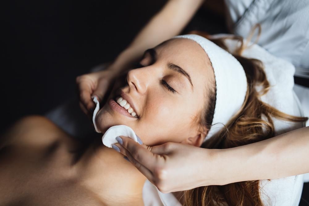 Woman Receiving Facial, Therapist Using Cotton Pads. She's Smiling, Wearing A Headband, At A Spa