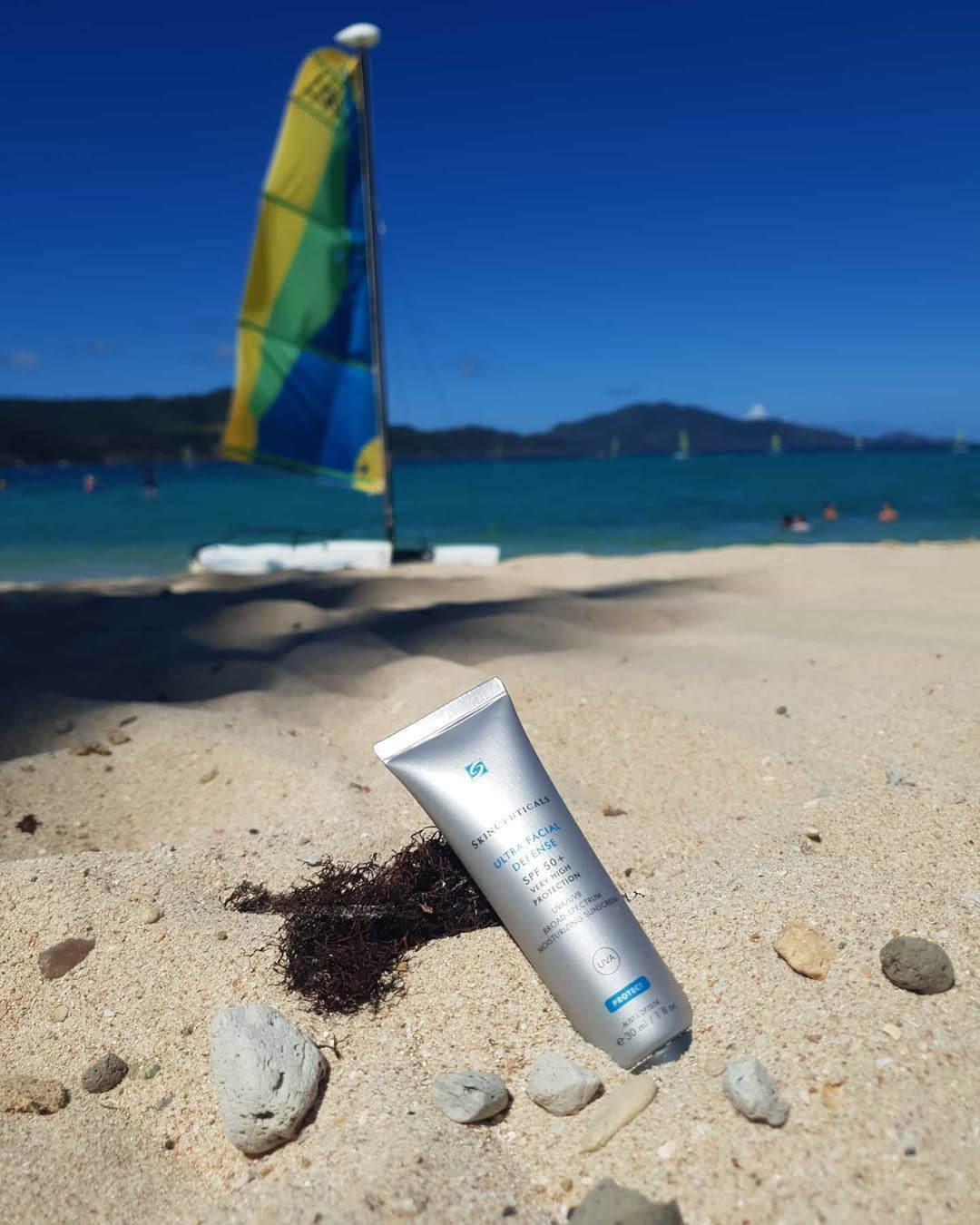 Silver Sunscreen Tube on Sandy Beach with Colourful Sailboat and Blue Water in The Background — Mackay Skin Clinic In North Mackay, QLD