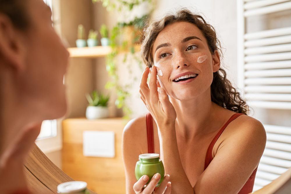 A Woman Is Applying Cream To Her Face In Front Of A Mirror — Mackay Skin Clinic In North Mackay, QLD