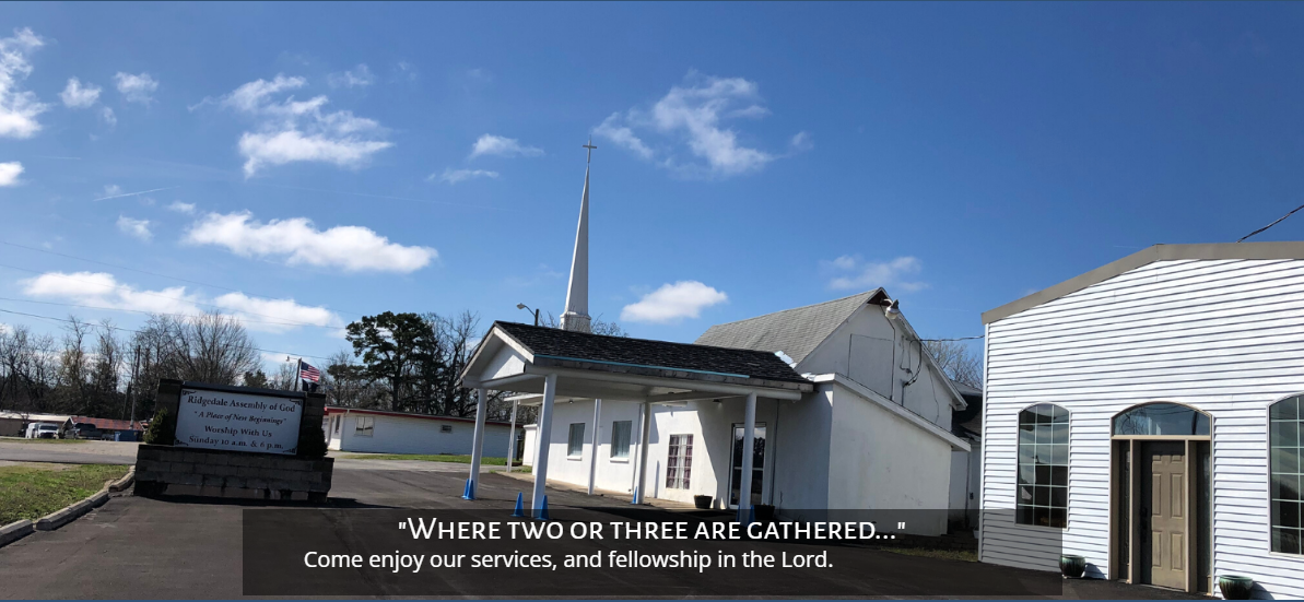 White church building with steeple and sign under a blue sky, seen from the street.