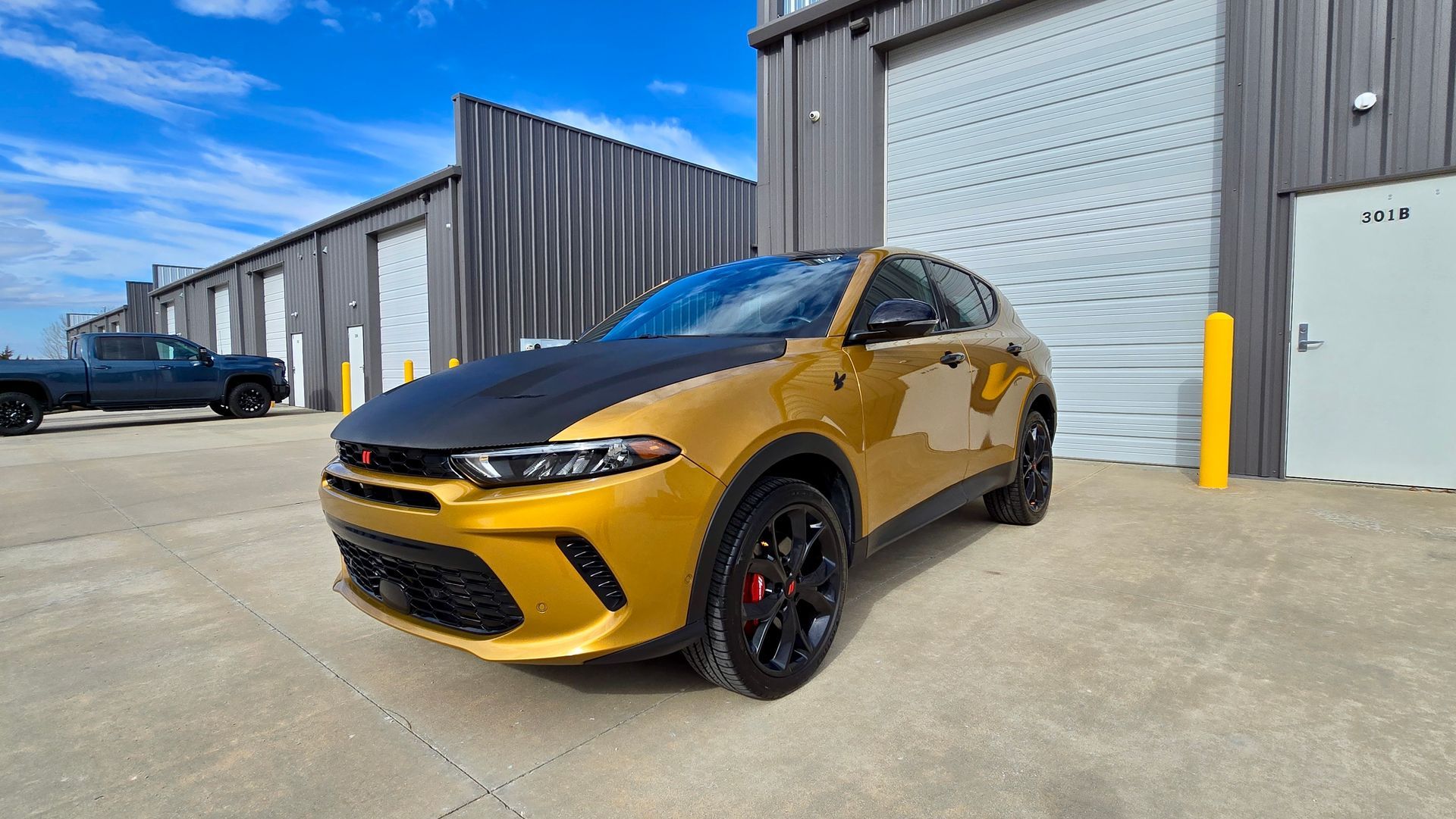 Golden Dodge Hornet with black hood parked outside a gray building on a sunny day.