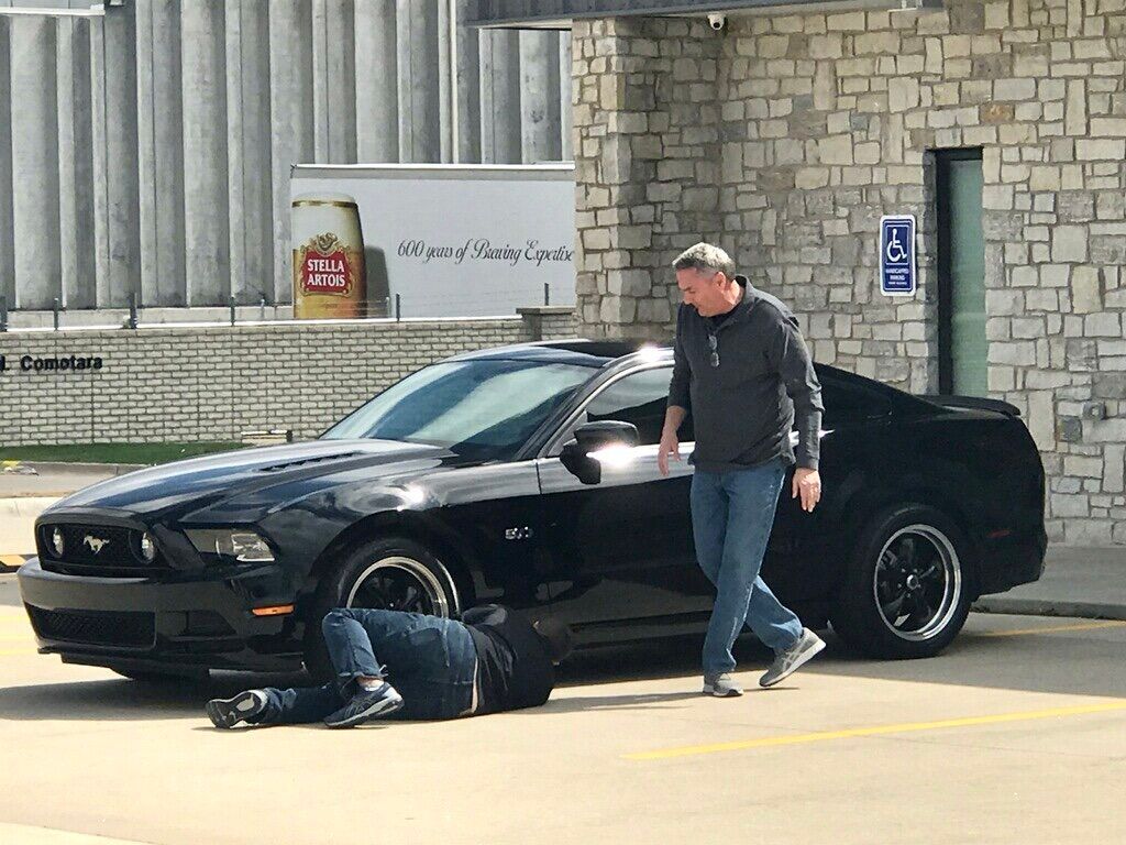 A man standing next to a black car with a man laying on the ground in front of it.