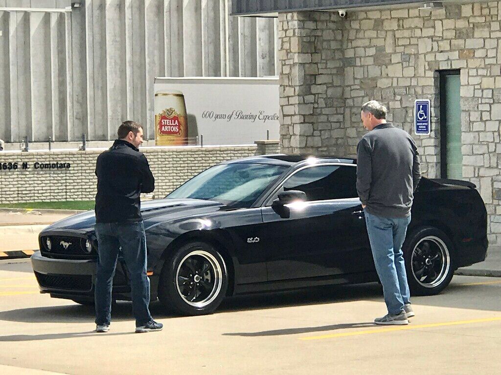 Two men are standing next to a black car in a parking lot.