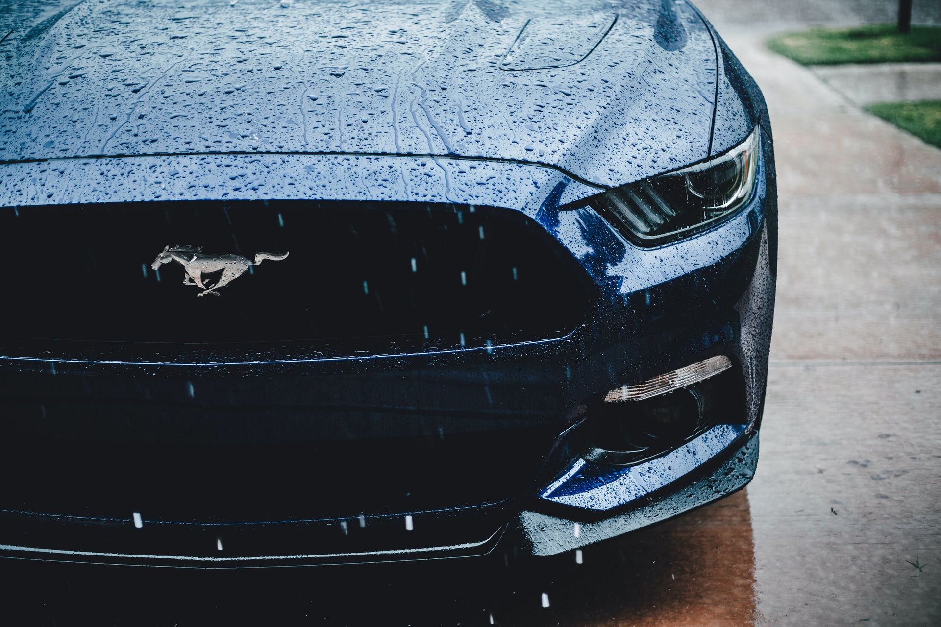 A blue mustang is parked in the rain on a sidewalk.