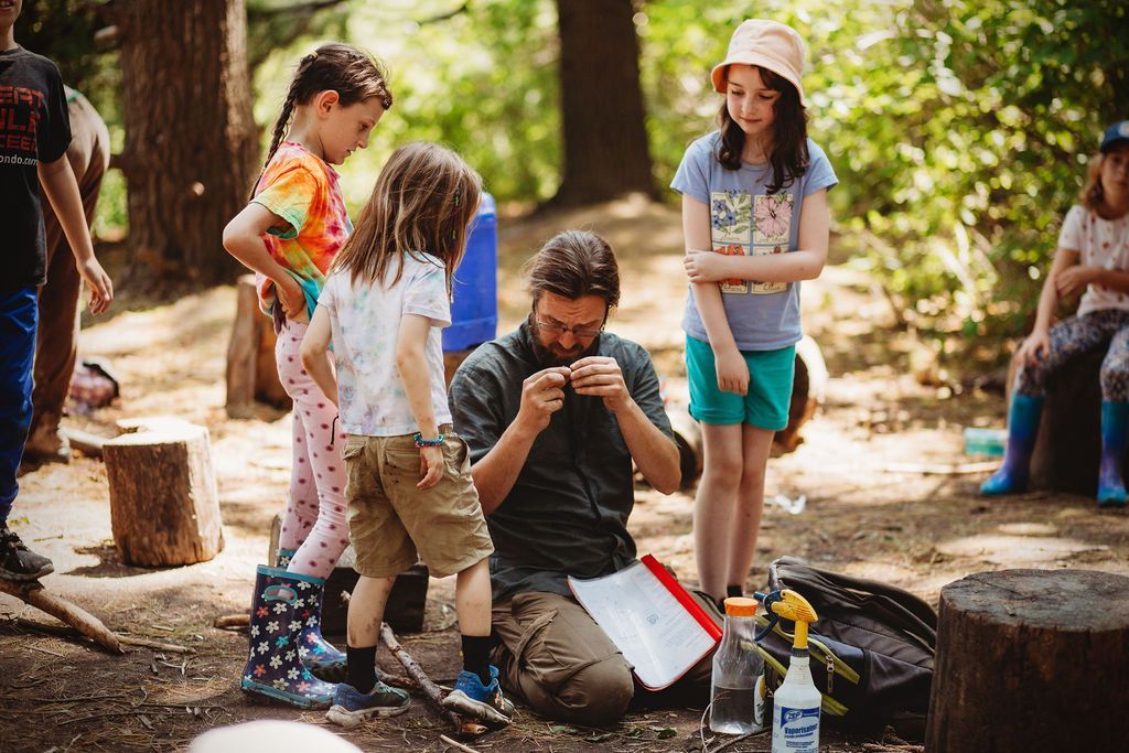 A man is kneeling down in the woods with a group of children.