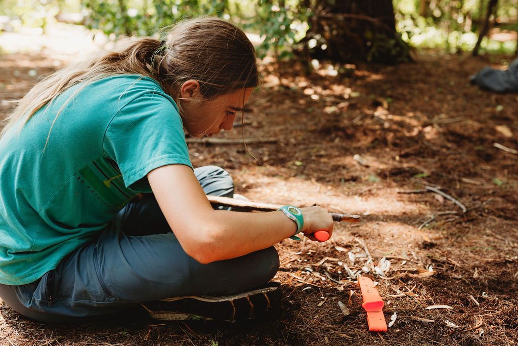 A woman is sitting on the ground in the woods cutting a piece of wood.