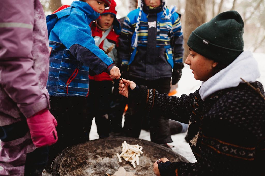 A woman is teaching a group of children how to make a fire in the snow.