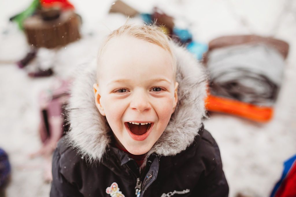 A young boy wearing a black jacket with a fur hood is laughing in the snow.
