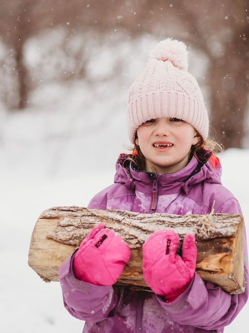 A little girl in a purple jacket and pink mittens is holding a log in the snow.