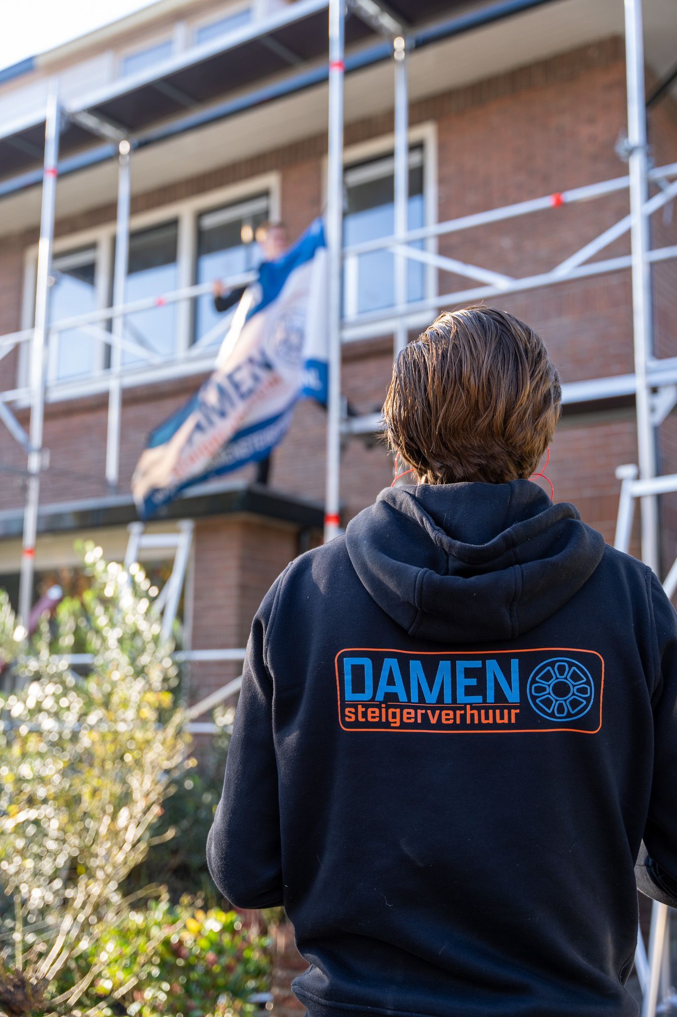 Een man in een zwarte hoodie staat voor een gebouw met een steiger.