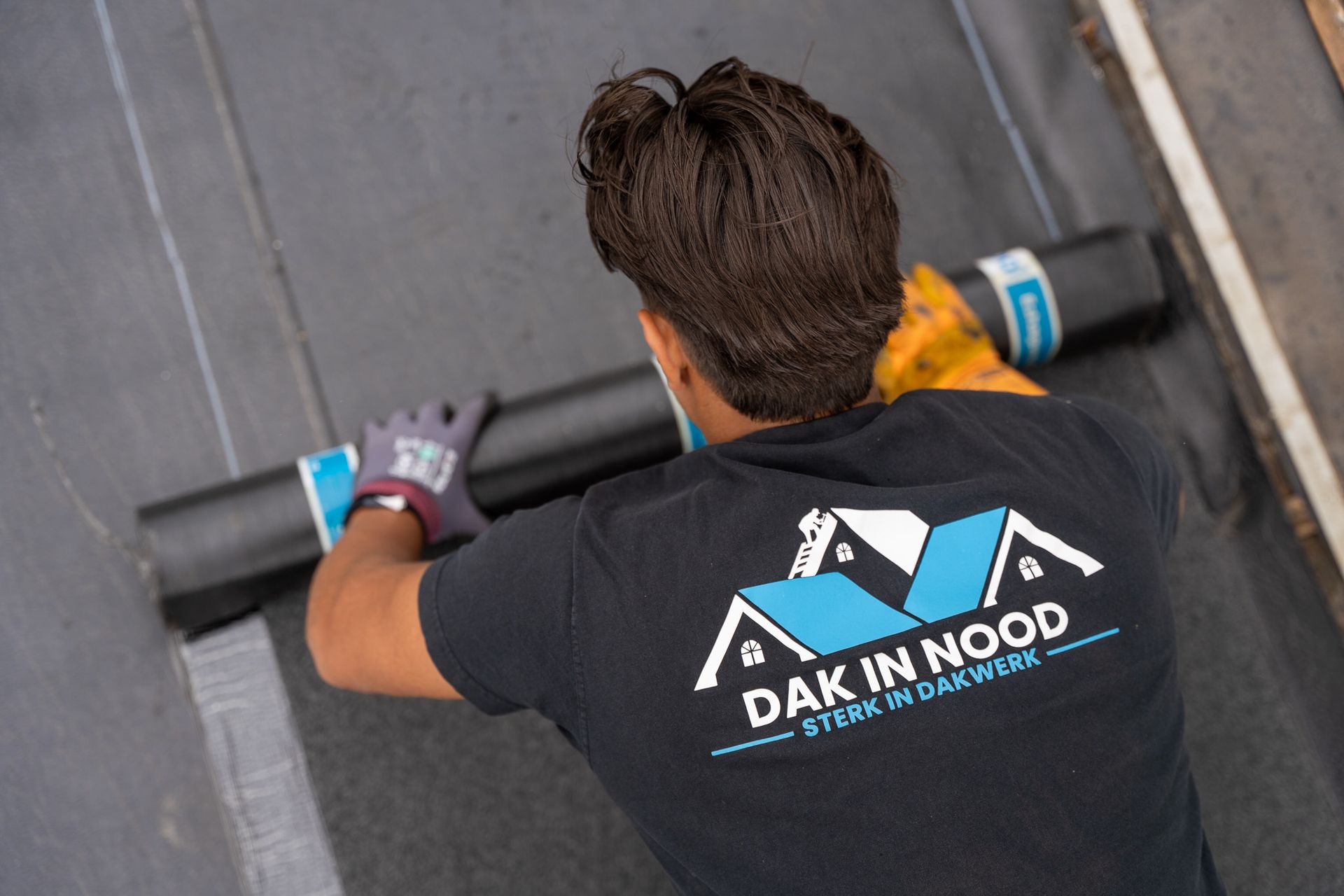 Een man met een zwart shirt met de tekst 'dak in nood' is op een dak aan het werk.
