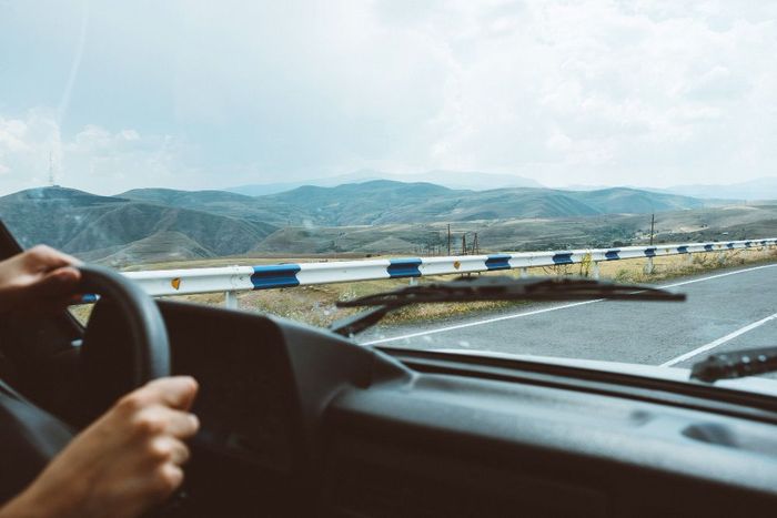 Hands gripping a steering wheel, driving on a road with mountains in the background under a cloudy sky.