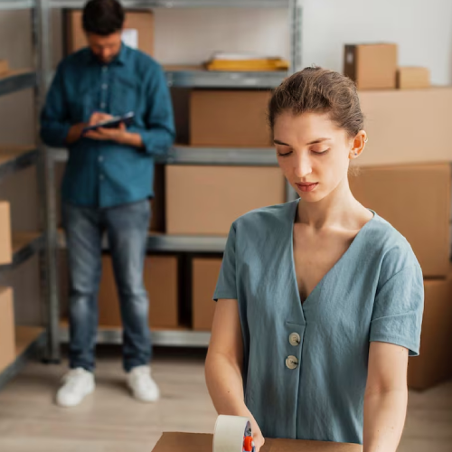Woman sealing a cardboard box in a warehouse. Man in background takes inventory. Shelves filled with boxes.