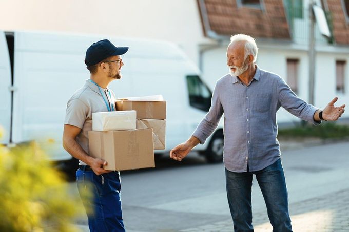 Delivery person handing packages to a man; white van in the background. Man gestures with surprise. Outdoors.