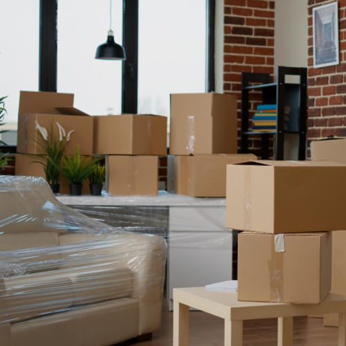 Room filled with moving boxes and furniture wrapped in plastic, near a window.