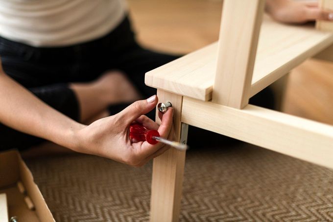 Person assembling wooden chair with a screwdriver indoors on a rug.