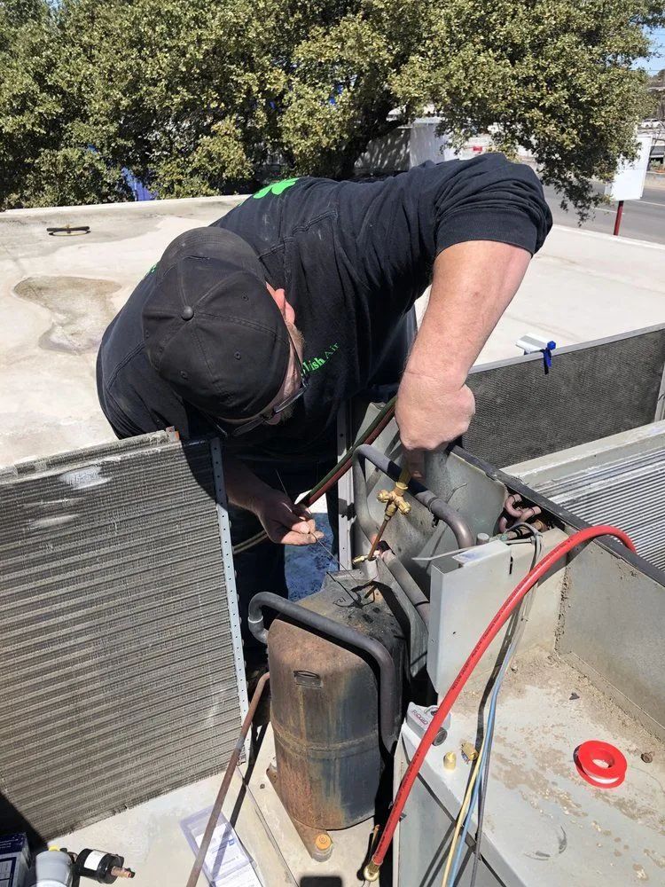 Technician working on rooftop HVAC unit, bent over with tools and hoses attached