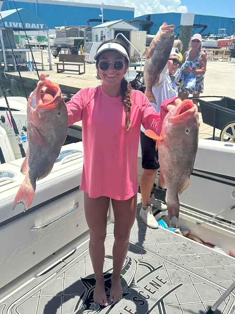 Woman on a boat holds up two large red fish she caught, smiling. Other people stand in the background.