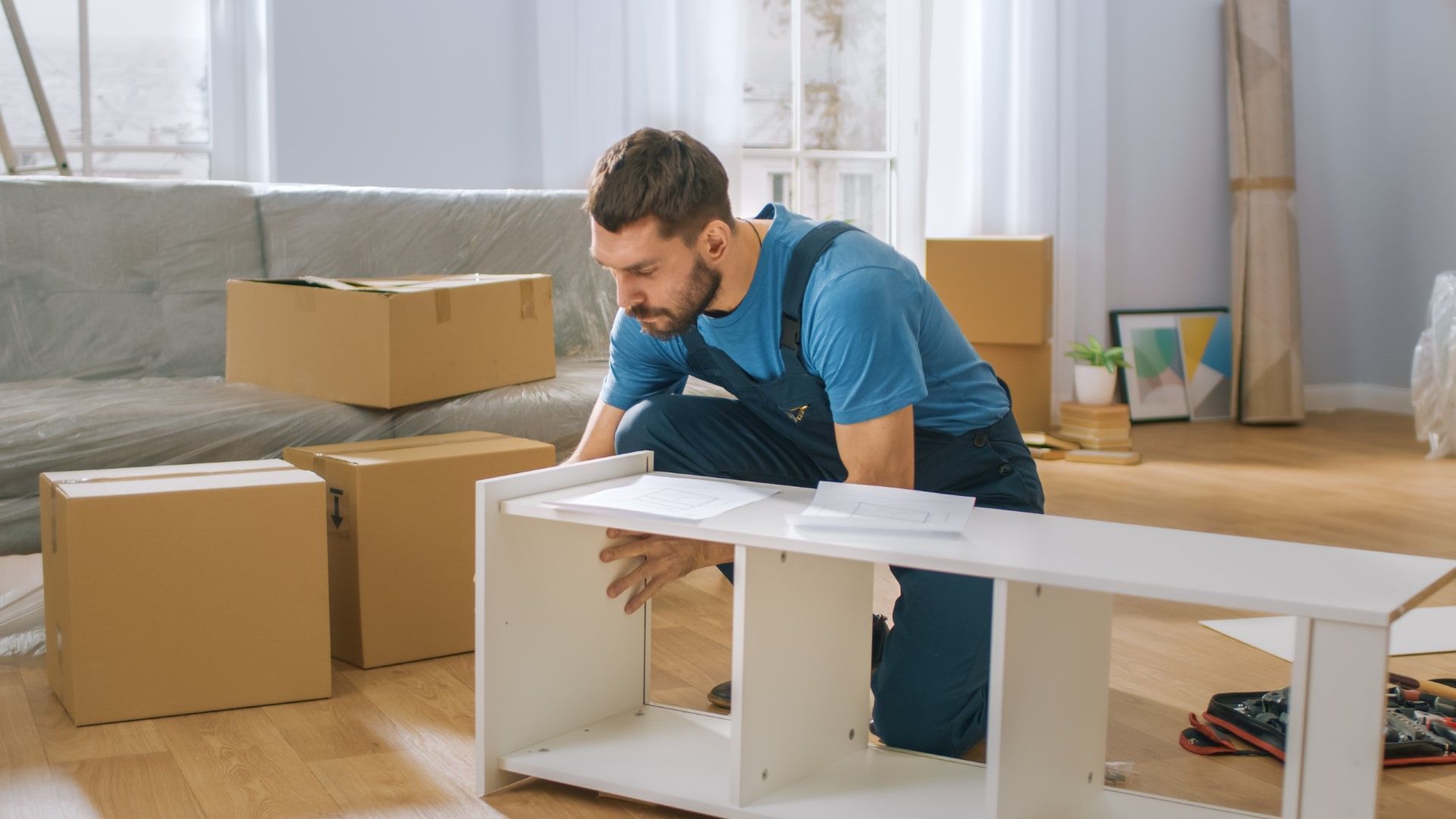 Man kneeling on a floor, assembling a white shelving unit among moving boxes in a bright room