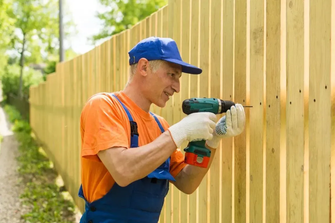 Worker in orange shirt drilling a wooden fence outdoors.