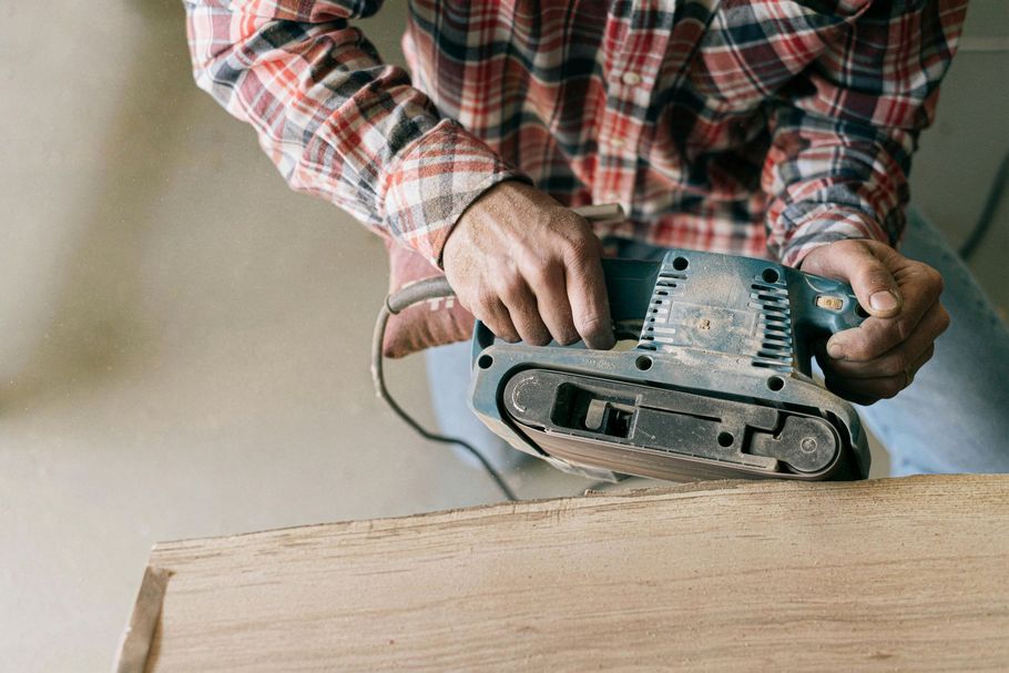 Person sanding a wooden surface with a handheld electric sander, wearing a red plaid shirt.