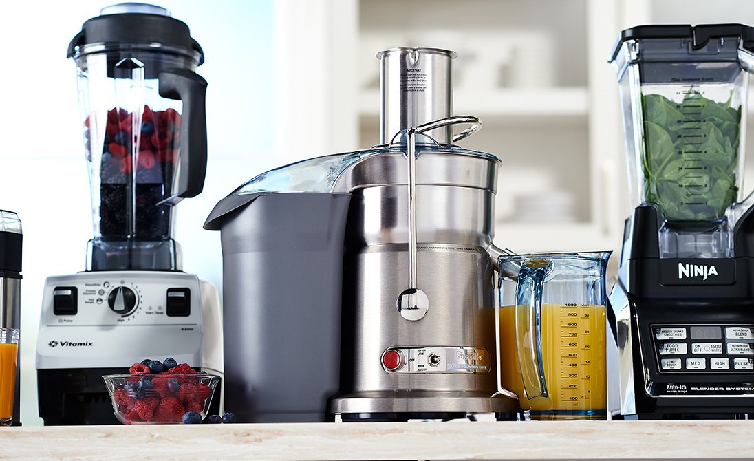 Juicers and blenders lined up on a counter, some with fruit and juice.