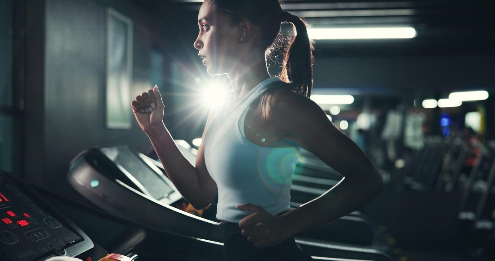 Woman running on a treadmill in a dimly lit gym, bright light behind her.