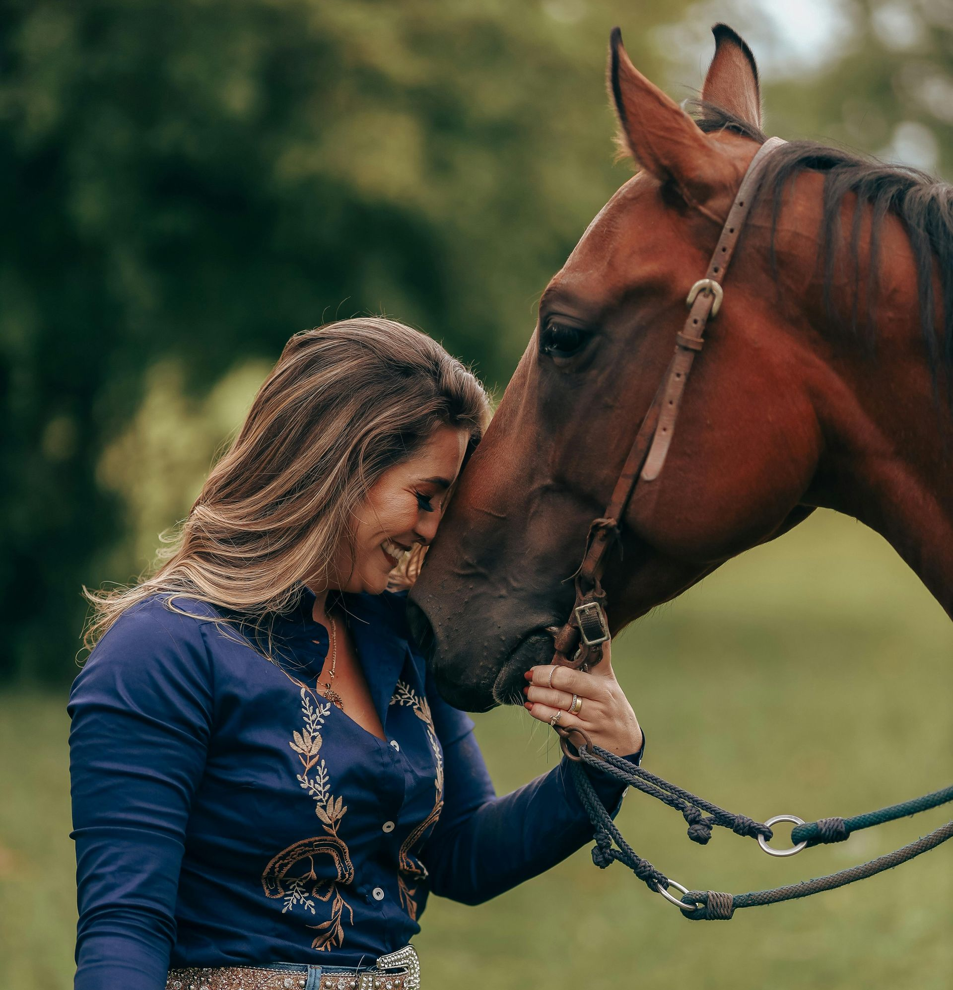 Woman in blue shirt lovingly touches a brown horse's head in a grassy field.