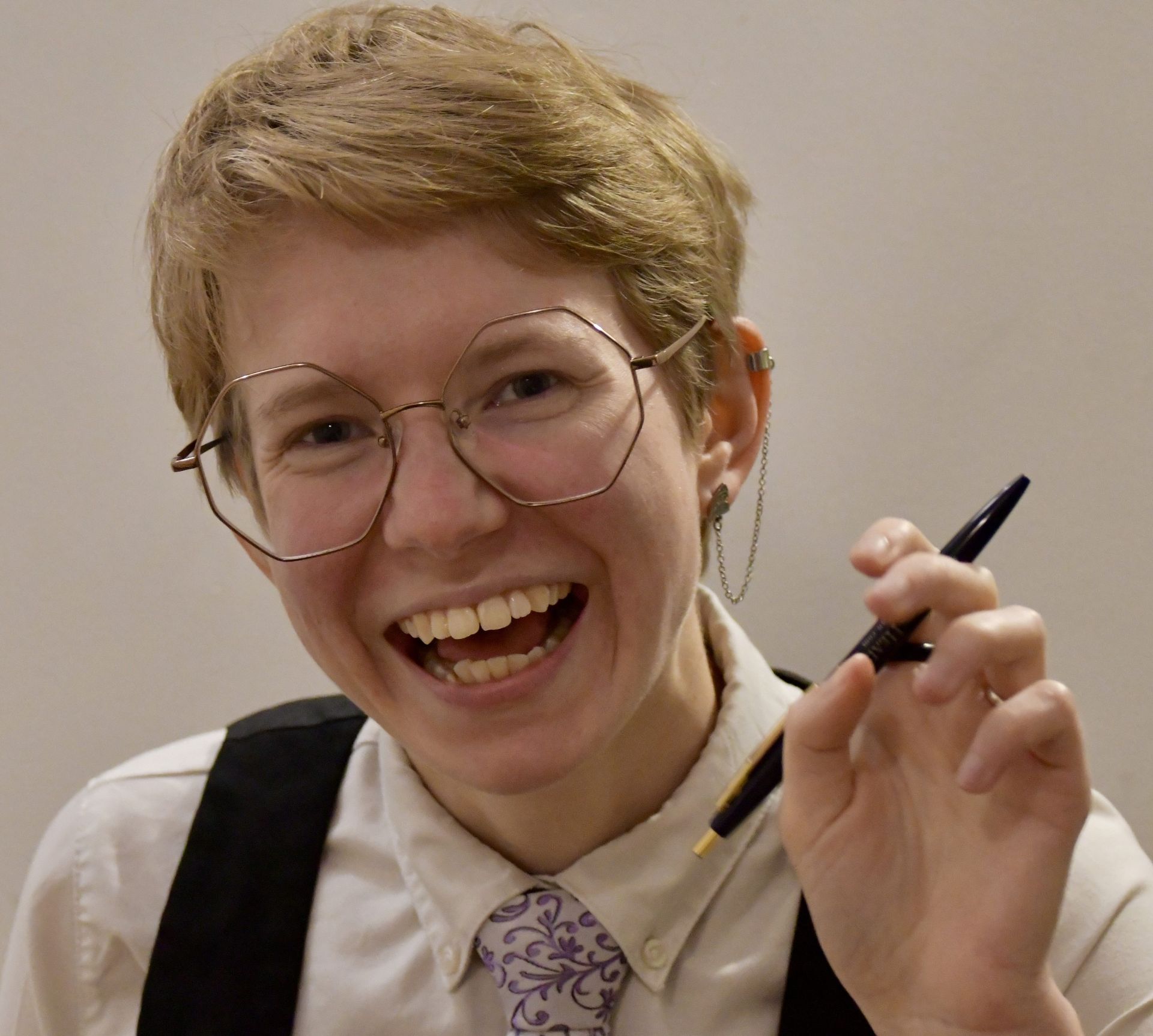 Photo of Alex Kingsley, who is a Caucasian person with short blond hair, gold wire-framed glasses and a big smile. They are holding a pen.