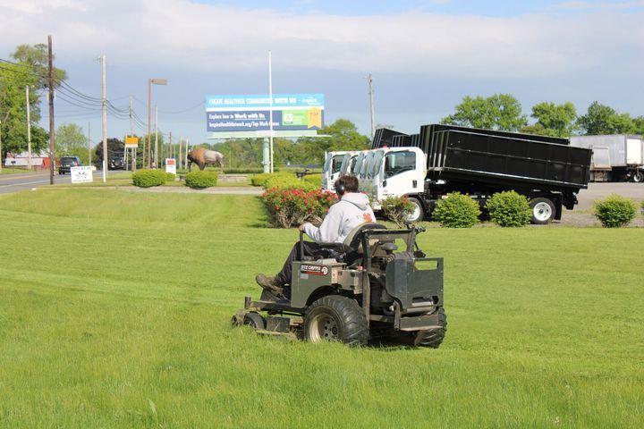 Man mowing grass with a riding mower. A truck, billboard, and road are in the background.
