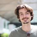 Un jeune homme avec une barbe et des lunettes de soleil sourit à la caméra.